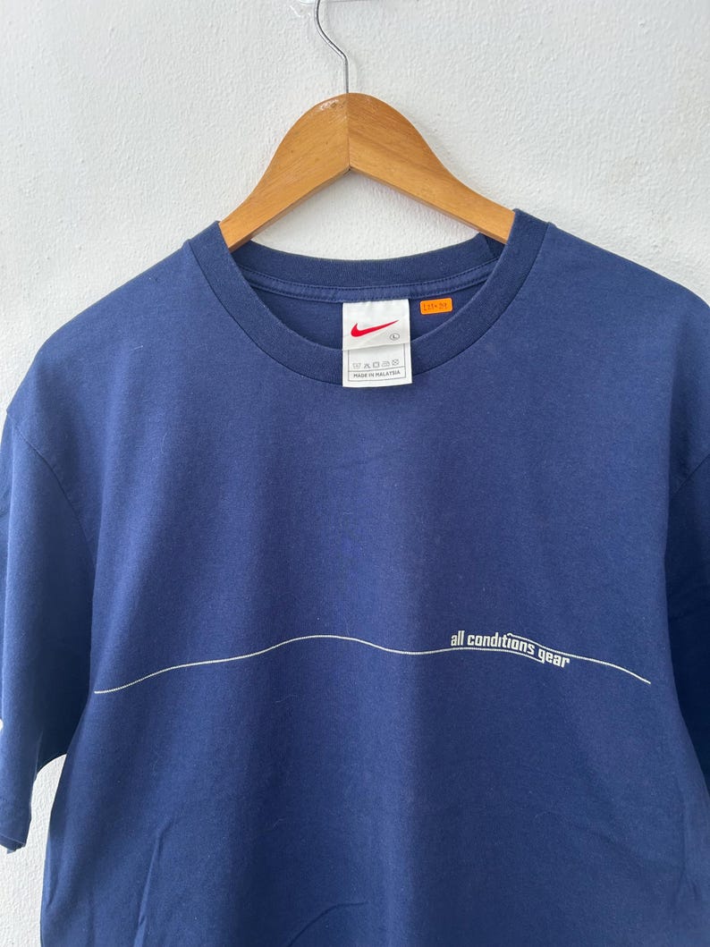May include: Navy blue t-shirt with a white horizontal line and the text "all conditions gear". A small Nike logo is visible on the tag. The shirt is hanging on a wooden hanger.