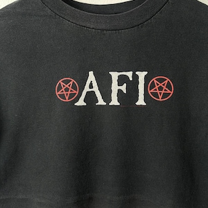 May include: A black crewneck t-shirt featuring the white text AFI centered on the chest. The letters are flanked on both sides by a red pentagram symbol. The shirt has a ribbed collar and is displayed on a wooden hanger against a plain background.