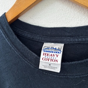 May include: Close-up of a black Gildan Activewear Heavy Weight Cotton t-shirt. The tag reads "M" and "100% Cotton Preshrunk Made in Nicaragua". The shirt is hanging on a wooden hanger against a white background.