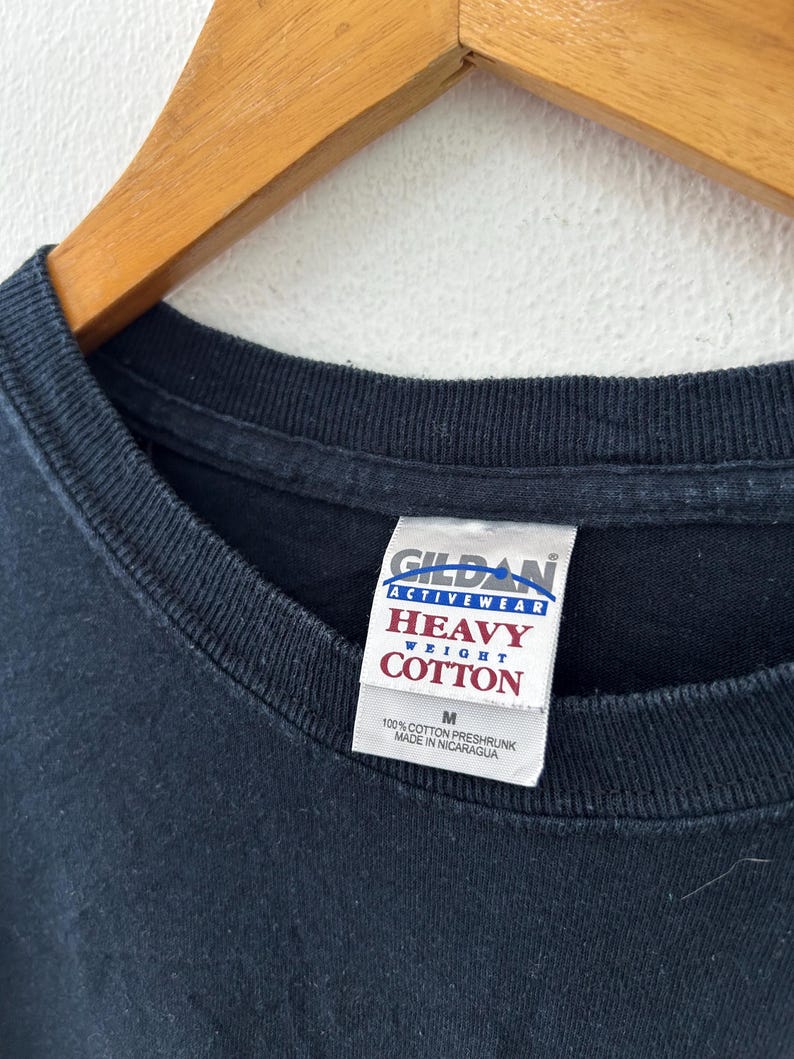 May include: Close-up of a black Gildan Activewear Heavy Weight Cotton t-shirt. The tag reads "M" and "100% Cotton Preshrunk Made in Nicaragua". The shirt is hanging on a wooden hanger against a white background.