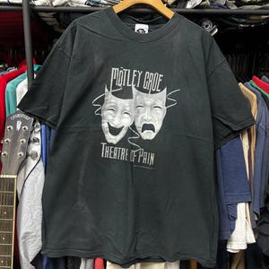 以下が含まれることがあります： 白いマスク2つと「Motley Crue Theatre of Pain」の文字が書かれた黒いTシャツ。