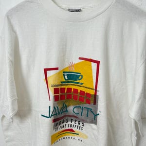 Puede incluir: Camiseta blanca con un diseño gráfico retro. El diseño presenta una taza de café, las palabras "JAVA CITY" y "ROASTERS OF FINE COFFEES, SACRAMENTO, CA." en un estilo colorido. La camiseta tiene cuello redondo y mangas cortas.