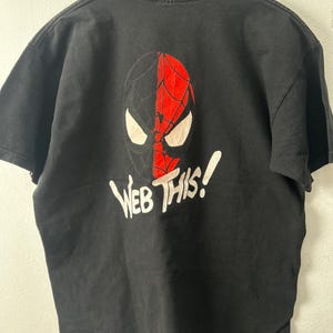 May include: A black short-sleeve t-shirt featuring a graphic of a split-colored mask in black and red with white eyes. Below the mask, the text "WEB THIS!" is printed in white lettering. The shirt is displayed on a wooden hanger.