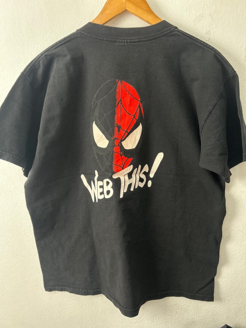 May include: A black short-sleeve t-shirt featuring a graphic of a split-colored mask in black and red with white eyes. Below the mask, the text "WEB THIS!" is printed in white lettering. The shirt is displayed on a wooden hanger.