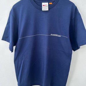 May include: Navy blue t-shirt with a white horizontal line and the text "all conditions gear". The shirt has a crew neck and short sleeves. A Nike logo and tag are visible near the neckline. The shirt is hanging on a wooden hanger.