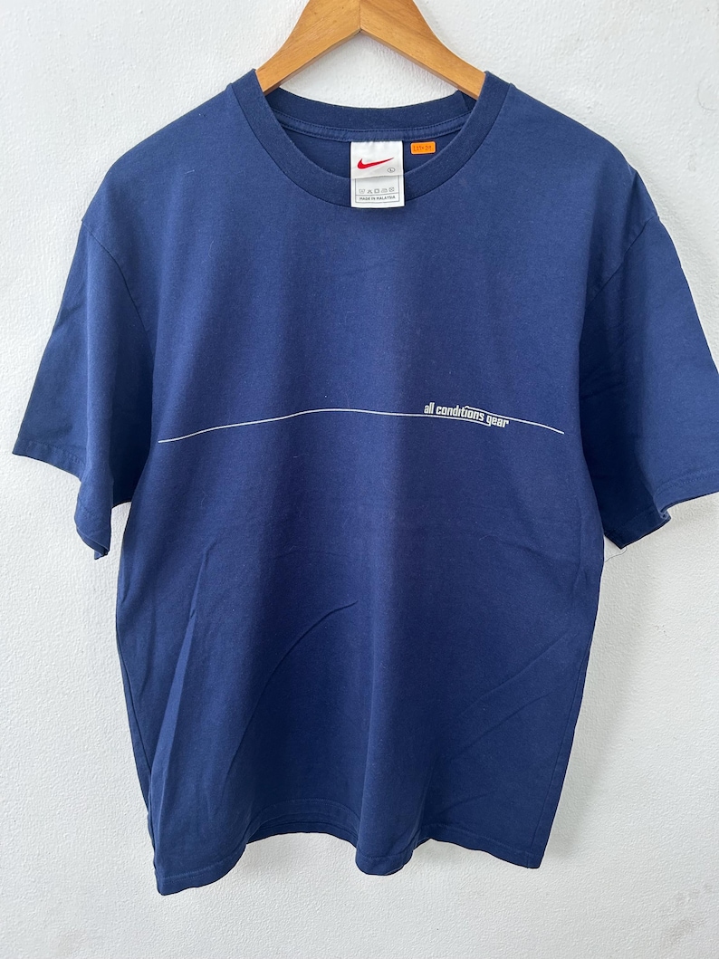 May include: Navy blue t-shirt with a white horizontal line and the text "all conditions gear". The shirt has a crew neck and short sleeves. A Nike logo and tag are visible near the neckline. The shirt is hanging on a wooden hanger.
