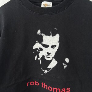 May include: Black crew neck t-shirt featuring a white and black image of a person's face. The name "rob thomas" is embroidered in red at the bottom of the shirt. The shirt has a kangaroo tag.