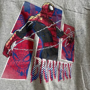 Puede incluir: Una camiseta gris con un estampado gr&aacute;fico rojo y azul de Spiderman. El gr&aacute;fico presenta a Spiderman en una telara&ntilde;a, con el texto "BRAVERY" impreso debajo.