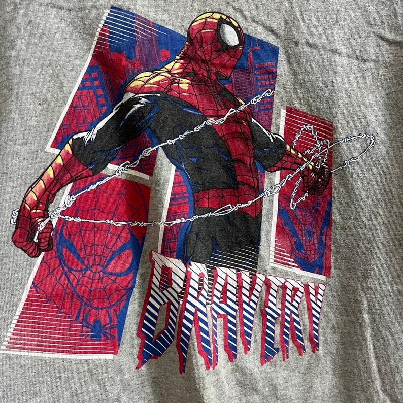 Puede incluir: Una camiseta gris con un estampado gr&aacute;fico rojo y azul de Spiderman. El gr&aacute;fico presenta a Spiderman en una telara&ntilde;a, con el texto "BRAVERY" impreso debajo.