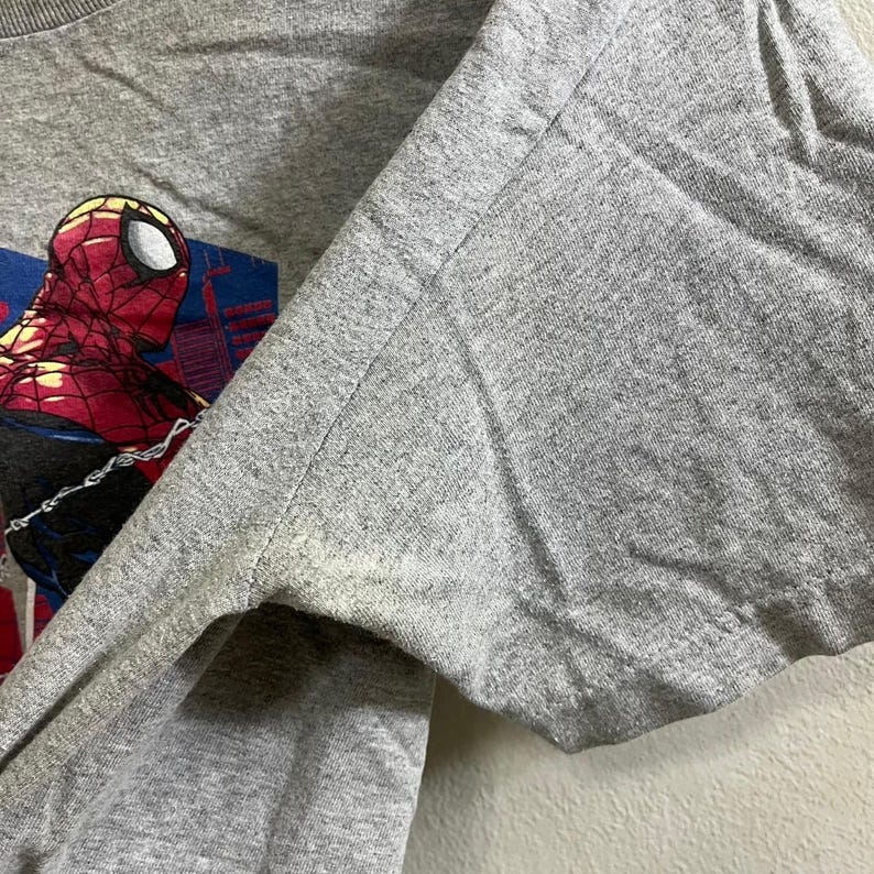 Puede incluir: Una camiseta gris con un estampado gr&aacute;fico de Spiderman balance&aacute;ndose en una telara&ntilde;a. El gr&aacute;fico es en rojo, azul y negro.