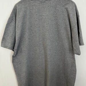 Puede incluir: Una camiseta gris jaspeada, exhibida en una percha de madera. La camiseta de manga corta est&aacute; hecha de una tela suave y texturizada. La parte trasera de la camiseta es visible, mostrando el cuello y la forma general de la prenda.