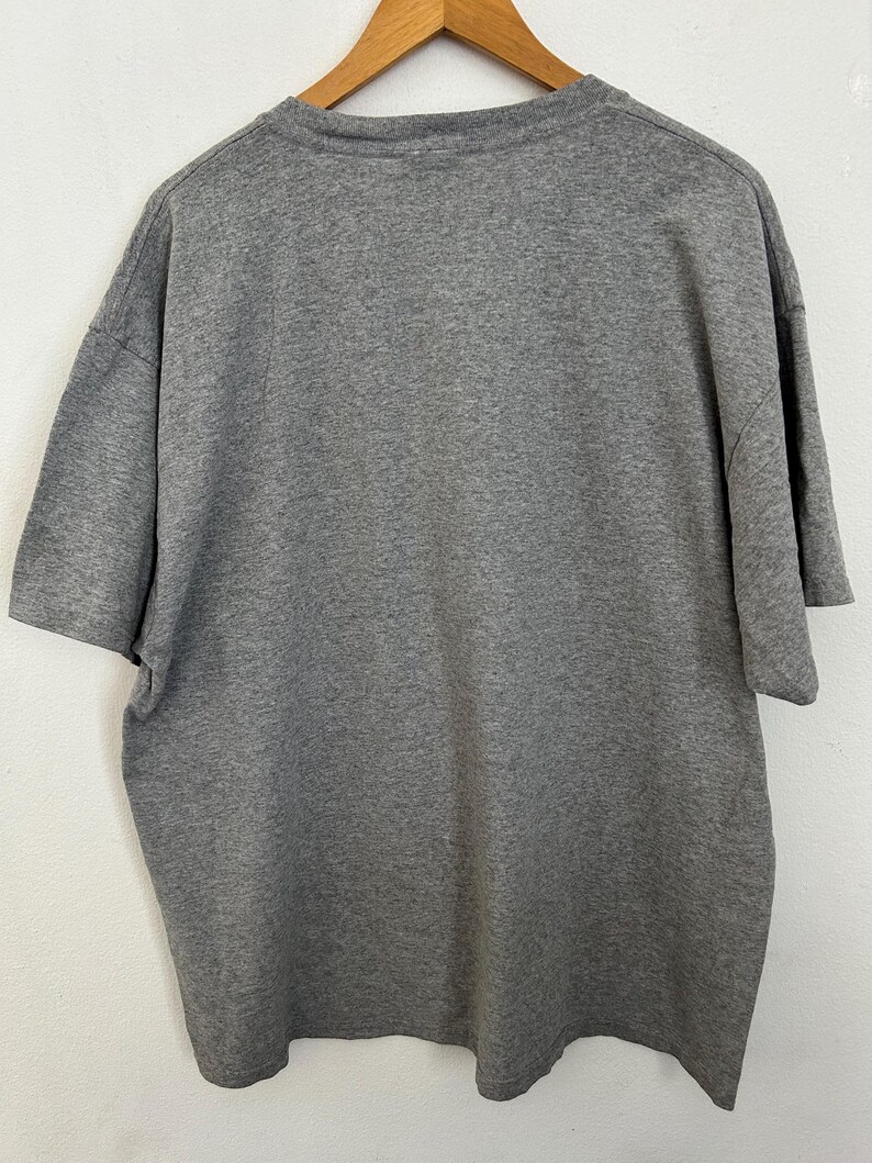 Puede incluir: Una camiseta gris jaspeada, exhibida en una percha de madera. La camiseta de manga corta est&aacute; hecha de una tela suave y texturizada. La parte trasera de la camiseta es visible, mostrando el cuello y la forma general de la prenda.