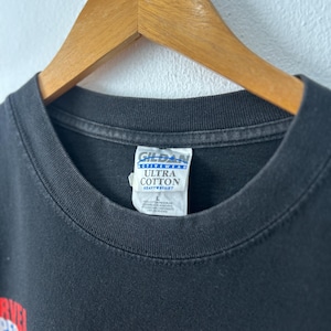 May include: A black t-shirt with a crew neckline, hanging on a wooden hanger. The interior neck label reads Gildan Activewear Ultra Cotton Heavyweight, with a size L indicated. A partial graphic with red and yellow text is visible on the lower left.