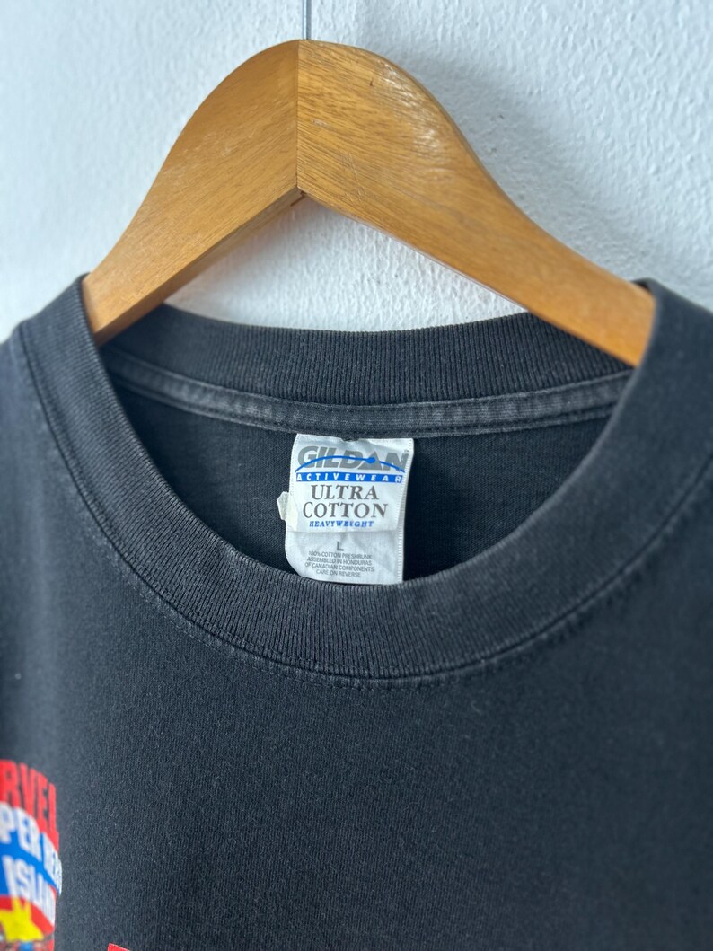 May include: A black t-shirt with a crew neckline, hanging on a wooden hanger. The interior neck label reads Gildan Activewear Ultra Cotton Heavyweight, with a size L indicated. A partial graphic with red and yellow text is visible on the lower left.