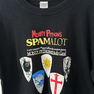 May include: A black t-shirt featuring the red and yellow text Monty Python's SPAMALOT, a new musical lovingly ripped off from the motion picture Monty Python and the Holy Grail, with five colourful shield graphics depicting a chicken, sun, tree, cross, and dragon.
