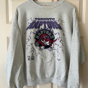 May include: A grey sweatshirt featuring a vintage Toronto Raptors logo. The logo depicts a purple and white "Toronto Raptors" text with a purple and white basketball dinosaur graphic. The sweatshirt has a crew neck and long sleeves.