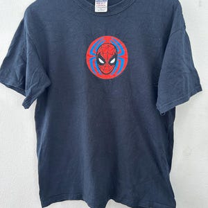 May include: A navy blue t-shirt featuring a Spider-Man graphic. The graphic is a circular design with a red spider web pattern, blue accents, and a black spider mask. The shirt is made of a soft cotton material.