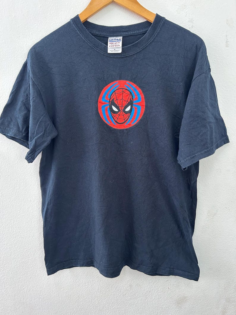 May include: A navy blue t-shirt featuring a Spider-Man graphic. The graphic is a circular design with a red spider web pattern, blue accents, and a black spider mask. The shirt is made of a soft cotton material.