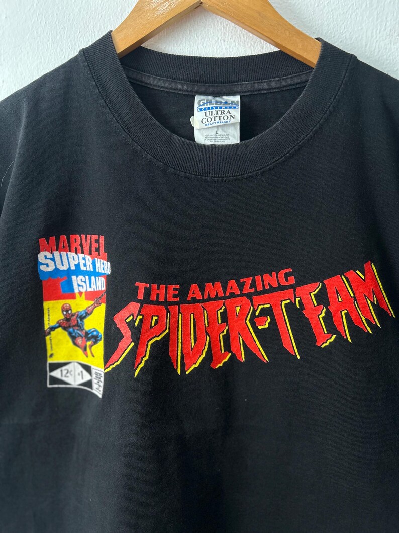 May include: A black t-shirt featuring a graphic of Spider-Man on a Marvel Super Hero Island comic book cover design. The text reads THE AMAZING SPIDER-TEAM in large red letters with yellow outlines. A Gildan Ultra Cotton size L tag is at the neck.
