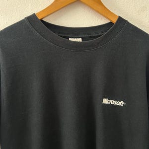 Może przedstawiać: Czarny t-shirt z okrągłym dekoltem z napisem "Microsoft" wydrukowanym na białym tle na górnej lewej piersi. Koszulka wisi na drewnianym wieszaku na neutralnym tle.