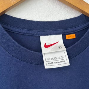 May include: Navy blue crew neck t-shirt with a white tag featuring a red Nike swoosh logo and the text "MADE IN MALAYSIA". The tag also includes washing instructions and the size "L".