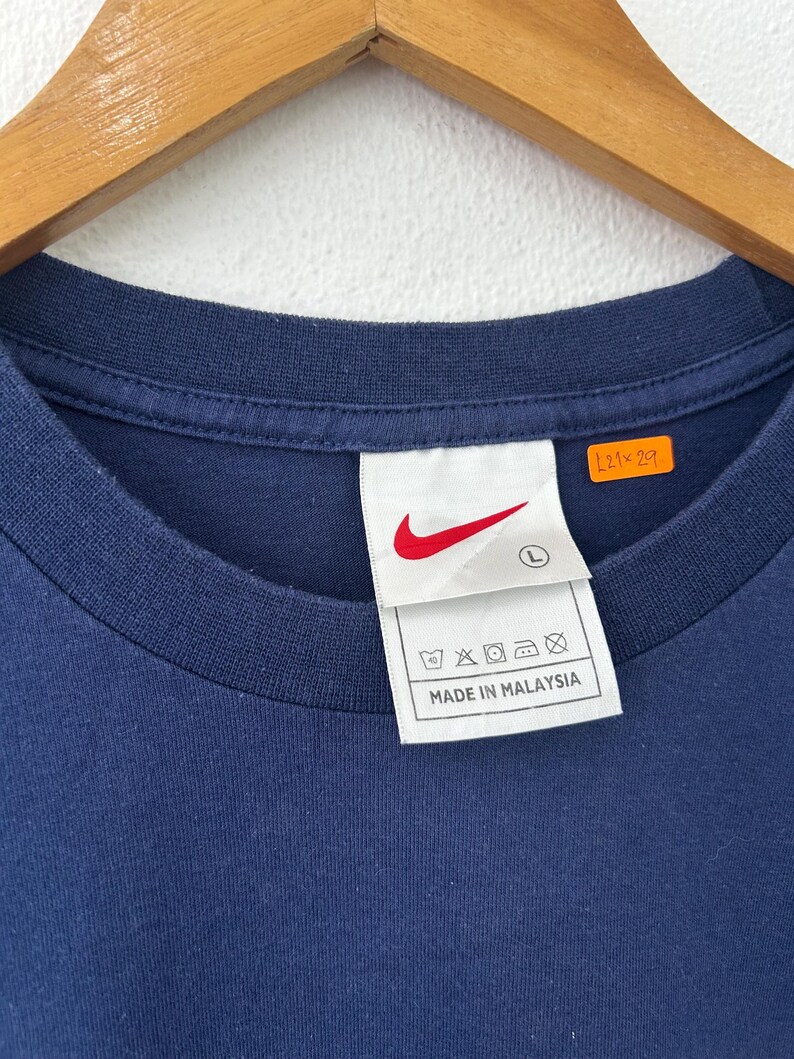 May include: Navy blue crew neck t-shirt with a white tag featuring a red Nike swoosh logo and the text "MADE IN MALAYSIA". The tag also includes washing instructions and the size "L".