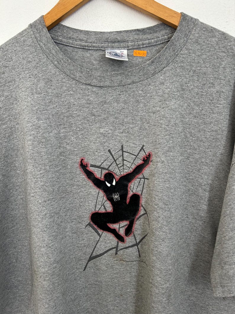 Puede incluir: Camiseta gris con un dise&ntilde;o de Spider-Man negro y detalles rojos, sobre un fondo de telara&ntilde;a gris. La camiseta tiene cuello redondo y mangas cortas. La etiqueta indica XL/30.