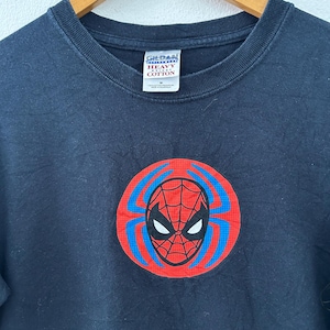 May include: A black t-shirt featuring a Spider-Man graphic. The graphic is a red circle with a blue web design and a Spider-Man mask in the center. The shirt is made of heavy cotton.