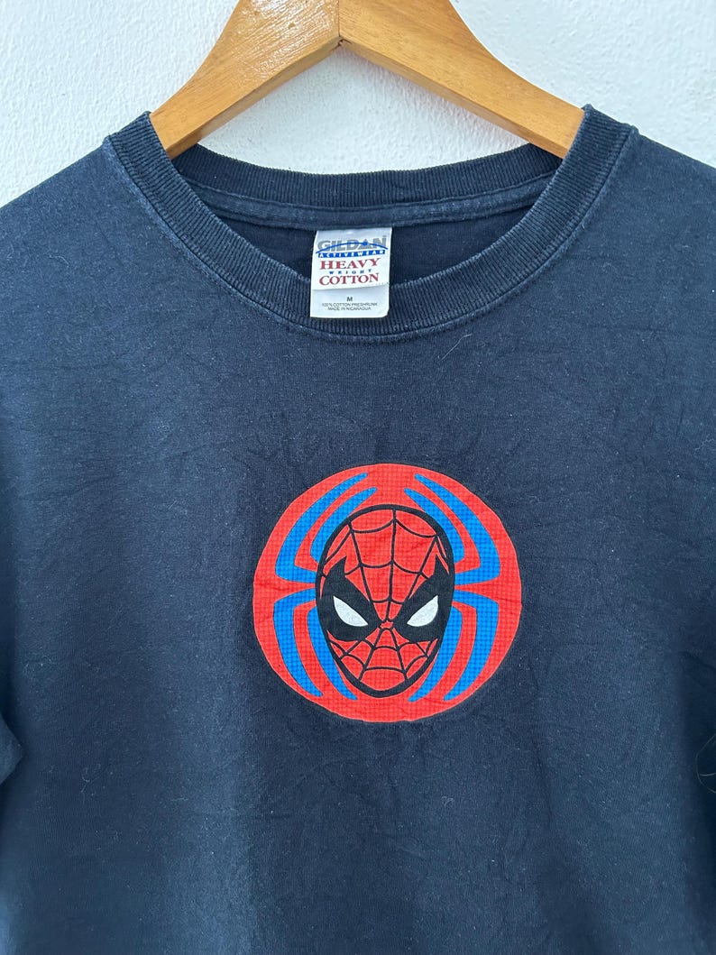 May include: A black t-shirt featuring a Spider-Man graphic. The graphic is a red circle with a blue web design and a Spider-Man mask in the center. The shirt is made of heavy cotton.