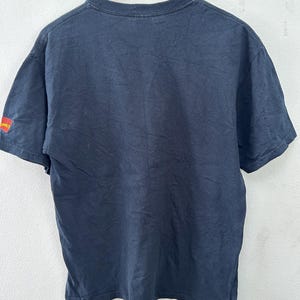 May include: A black t-shirt hanging on a wooden hanger. The shirt has short sleeves and a crew neck. The sleeve has a small red and yellow graphic that says "Alive Comics". The shirt is slightly wrinkled.
