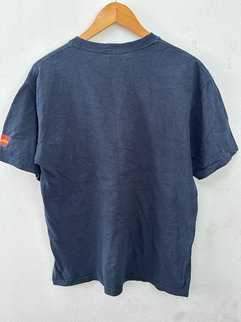 May include: A black t-shirt hanging on a wooden hanger. The shirt has short sleeves and a crew neck. The sleeve has a small red and yellow graphic that says "Alive Comics". The shirt is slightly wrinkled.