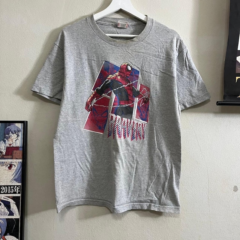 Puede incluir: Camiseta gris con un gr&aacute;fico de Spider-Man en un traje rojo y azul, con el texto "BRAVERY" impreso debajo.