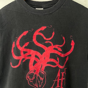 May include: Black t-shirt featuring a red graphic of intertwined snakes and a heart-shaped design. The text "AH" is printed in red below the graphic. The shirt is displayed on a wooden hanger.