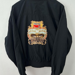 May include: Black bomber jacket with a ribbed collar and cuffs. The back features an embroidered design with the Pepsi logo and "Michael Jackson Dangerous World Tour" text. The jacket is hanging on a wooden hanger.