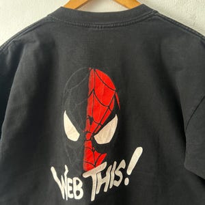 May include: A black short-sleeved t-shirt featuring a graphic of a split-colored mask, half black and half red with a web pattern. Below the mask, the text "WEB THIS!" is printed in white. The shirt is displayed on a wooden hanger.