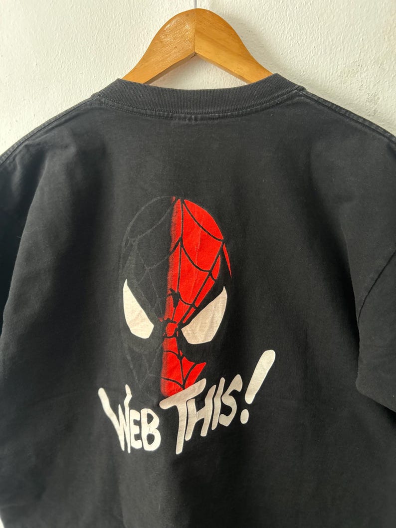 May include: A black short-sleeved t-shirt featuring a graphic of a split-colored mask, half black and half red with a web pattern. Below the mask, the text "WEB THIS!" is printed in white. The shirt is displayed on a wooden hanger.
