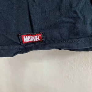 Op de afbeelding: Zwart T-shirt met een rood "MARVEL" label aan de onderkant.