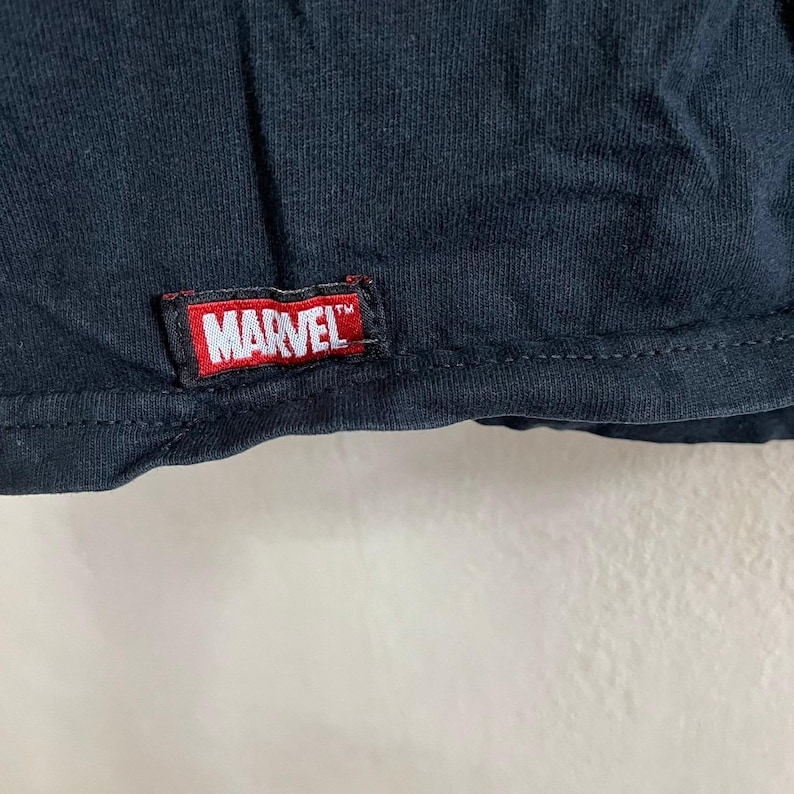 Op de afbeelding: Zwart T-shirt met een rood "MARVEL" label aan de onderkant.