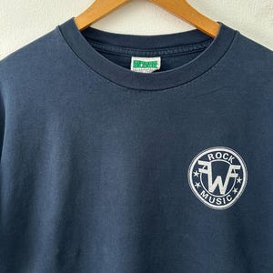 Könnte beinhalten: Marineblaues T-Shirt mit einem weißen kreisförmigen Logo mit der Aufschrift "ROCK MUSIC" und einem stilisierten "W" in der Mitte. Das T-Shirt hängt an einem Holzbügel.