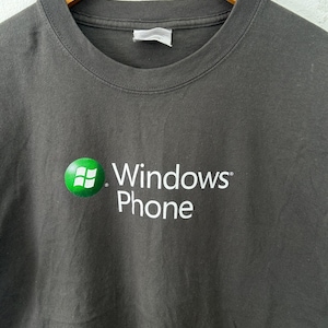 Op de afbeelding: Donkergrijs t-shirt met het Windows Phone-logo. Het logo is een groene cirkel met een wit rasterontwerp en de woorden "Windows Phone" zijn in het wit gedrukt.