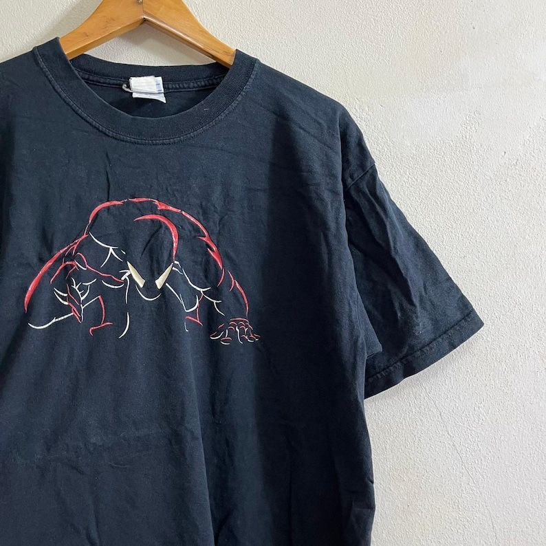 Op de afbeelding: Zwart T-shirt met een rode en witte afbeelding van een gehurkte figuur.