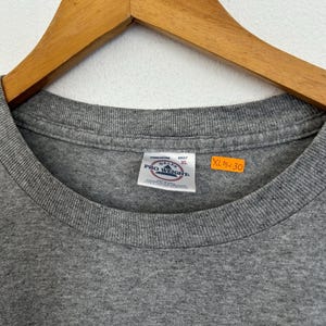 Puede incluir: Una camiseta gris con cuello redondo, colgada de una percha de madera. La camiseta tiene una etiqueta que dice "PRO WEIGHT" y "ADULT XL". Las medidas de la camiseta son XL 63.5cm x 76.2cm.