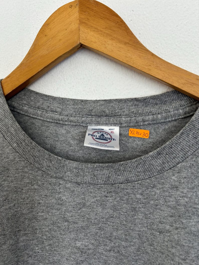 Puede incluir: Una camiseta gris con cuello redondo, colgada de una percha de madera. La camiseta tiene una etiqueta que dice "PRO WEIGHT" y "ADULT XL". Las medidas de la camiseta son XL 63.5cm x 76.2cm.