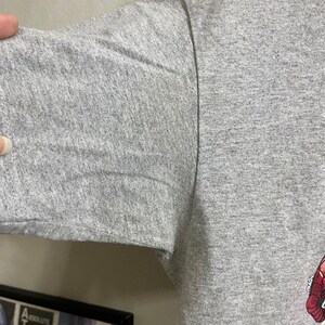 Puede incluir: Una camiseta gris con un estampado gr&aacute;fico descolorido de color rojo y negro. La camiseta est&aacute; hecha de un material suave y c&oacute;modo.