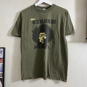 May include: A green t-shirt featuring a black and white image of Jimi Hendrix. The text "Jimi Hendrix" is printed in yellow above the image.