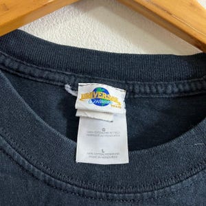 Op de afbeelding: Zwart T-shirt met een Universal Orlando logo-label. Het label is wit met blauwe tekst en een wereldbeeld. Het label zegt "Universal Orlando" en "THAD". Het label bevat ook maat- en wasinstructies.