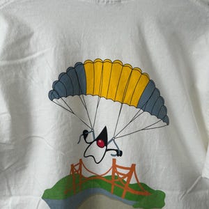 以下が含まれることがあります： 白いTシャツで、赤い円形の目を持つ様式化されたキャラクターが、黄色とダークグレーのパラシュートで、オレンジ色の橋がある緑の風景の上を降下しているグラフィックプリントが施されています。デザインは背面の中心に配置されています。