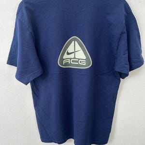 May include: Navy blue t-shirt with a large, light green and white ACG logo on the back. The logo features a Nike swoosh and the letters "ACG" in a sans-serif font. The shirt is made of a soft, likely cotton material.