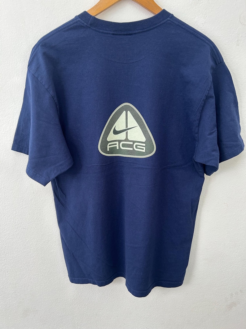 May include: Navy blue t-shirt with a large, light green and white ACG logo on the back. The logo features a Nike swoosh and the letters "ACG" in a sans-serif font. The shirt is made of a soft, likely cotton material.