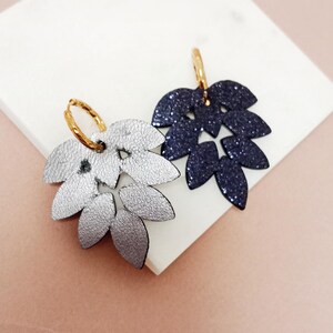 Pode incluir: Dois brincos em forma de folha, um prateado, outro azul marinho com glitter. Ambos têm fechos de argola dourados. Os brincos são exibidos em uma superfície branca sobre um fundo rosa. O brinco prateado tem um acabamento texturizado.